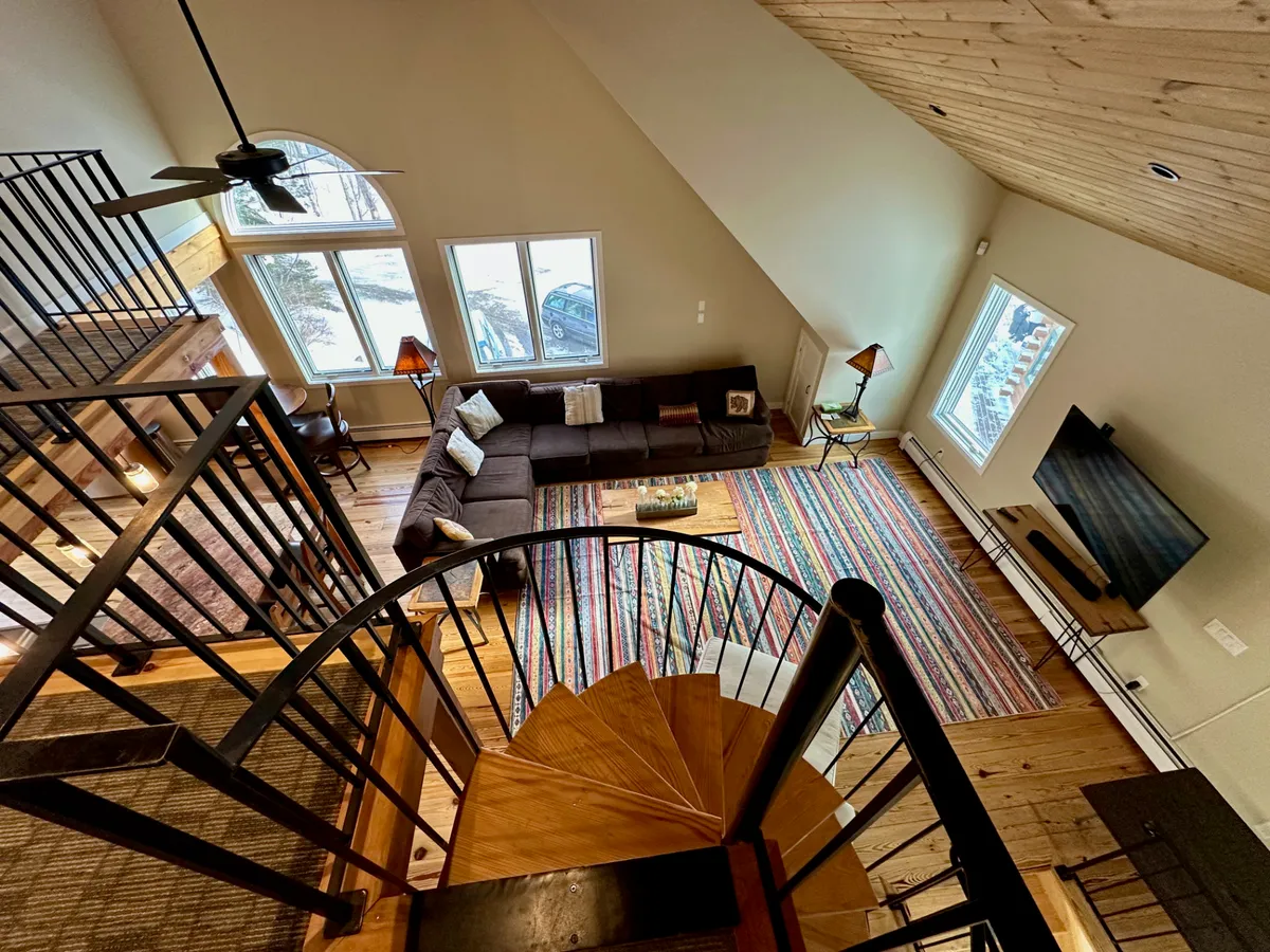 A cozy, inviting living room with a sectional sofa and colorful area rug set beneath large windows. The spiral staircase adds architectural interest, and the space is flooded with natural light. The area is perfect for relaxing with family, hosting gatherings, or watching movies, with a flat-screen TV visible in the background. Warm wooden accents and a fireplace create a comforting alpine ambiance.