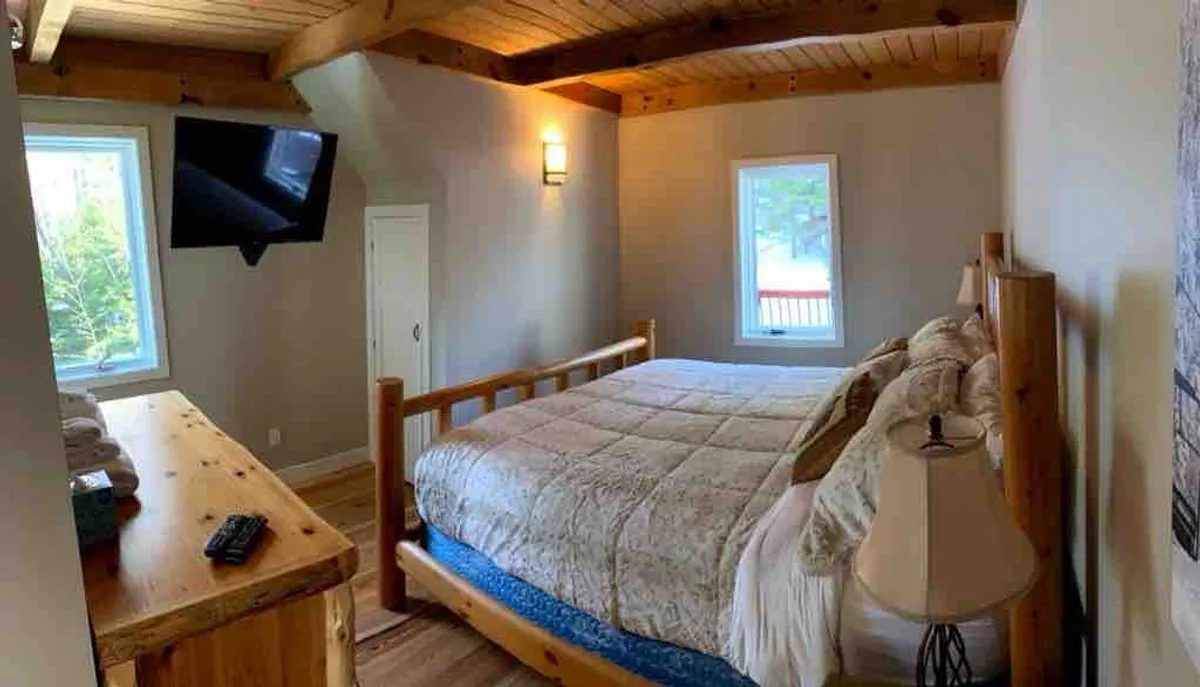 A cozy bedroom featuring a plush bed positioned near large windows that let in natural light. The space is accented with wooden furniture, a bedside lamp, and a TV, creating a restful and inviting atmosphere suitable for relaxation and comfort.