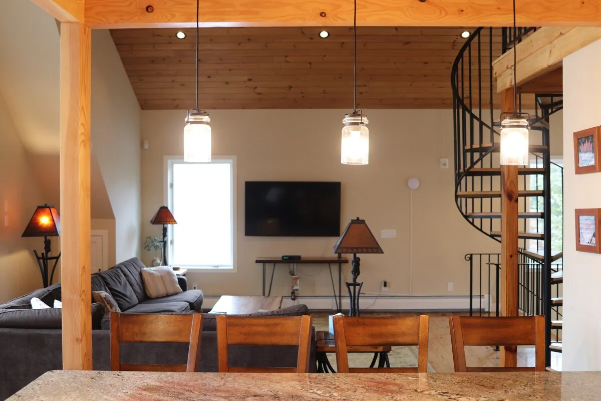 Spacious living area featuring a comfortable seating arrangement, a modern television setup, and a spiral staircase that leads to other parts of the home. The dining table is visible, and the design supports both entertainment and relaxation. Soft lighting and thoughtful decor create a warm, welcoming vibe, ideal for family gatherings or movie nights after a day on the slopes.