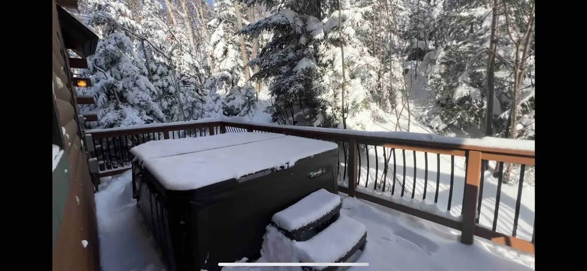 An exterior view of the snow-covered deck featuring a steaming hot tub surrounded by trees. The tranquil setting invites guests to soak under the stars after a day of skiing, with the natural landscape providing a gorgeous backdrop. The deck seamlessly connects outdoor relaxation to the comfort of the interior living space.