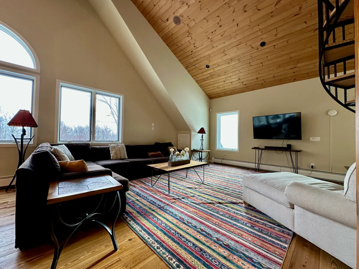 This image features a comfortable seating area with a sofa, coffee table, colorful rug, spiral staircase, and expansive windows that reveal views of the outdoors. The scene feels welcoming and encourages group gatherings, relaxation, and soaking in the natural vistas.