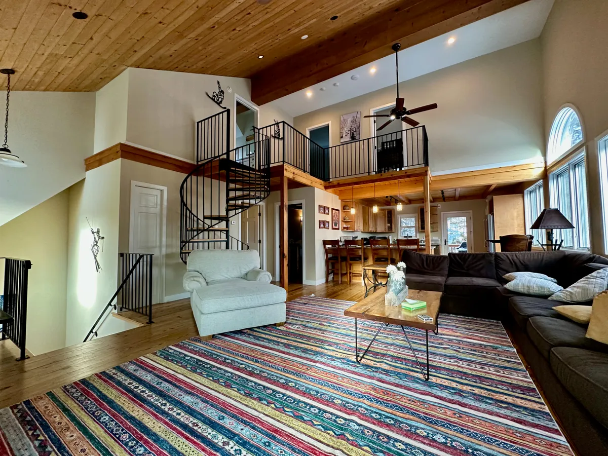 The image showcases the stylish and inviting living area of the guesthouse, featuring a striking spiral staircase, a vibrant area rug that adds warmth, expansive windows letting in natural light and mountain views, and a charming wooden ceiling that exudes rustic elegance. The space is arranged for both relaxation and togetherness, perfect for gathering with family or friends after a day on the slopes.