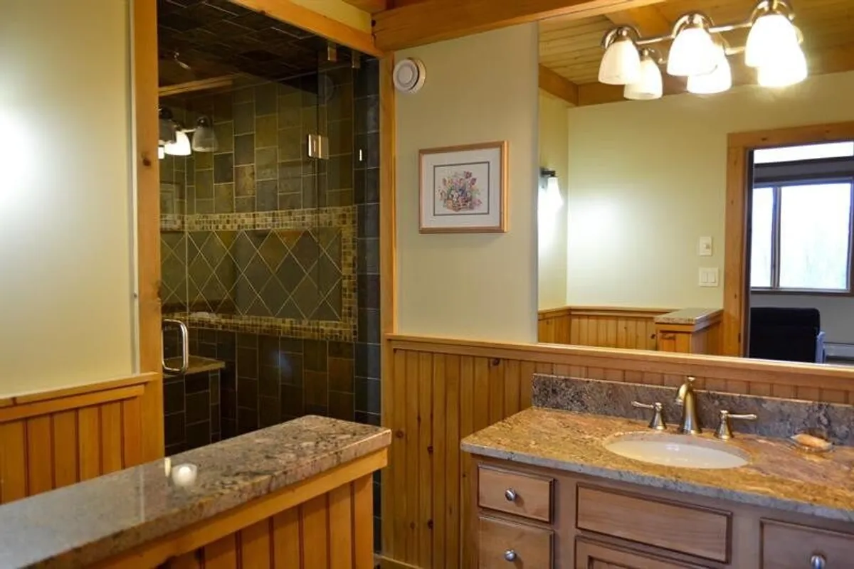 A modern bathroom with a granite countertop, glass shower, wood paneling, and framed artwork. The design combines luxury and warmth, creating a calming space for relaxation and self-care.