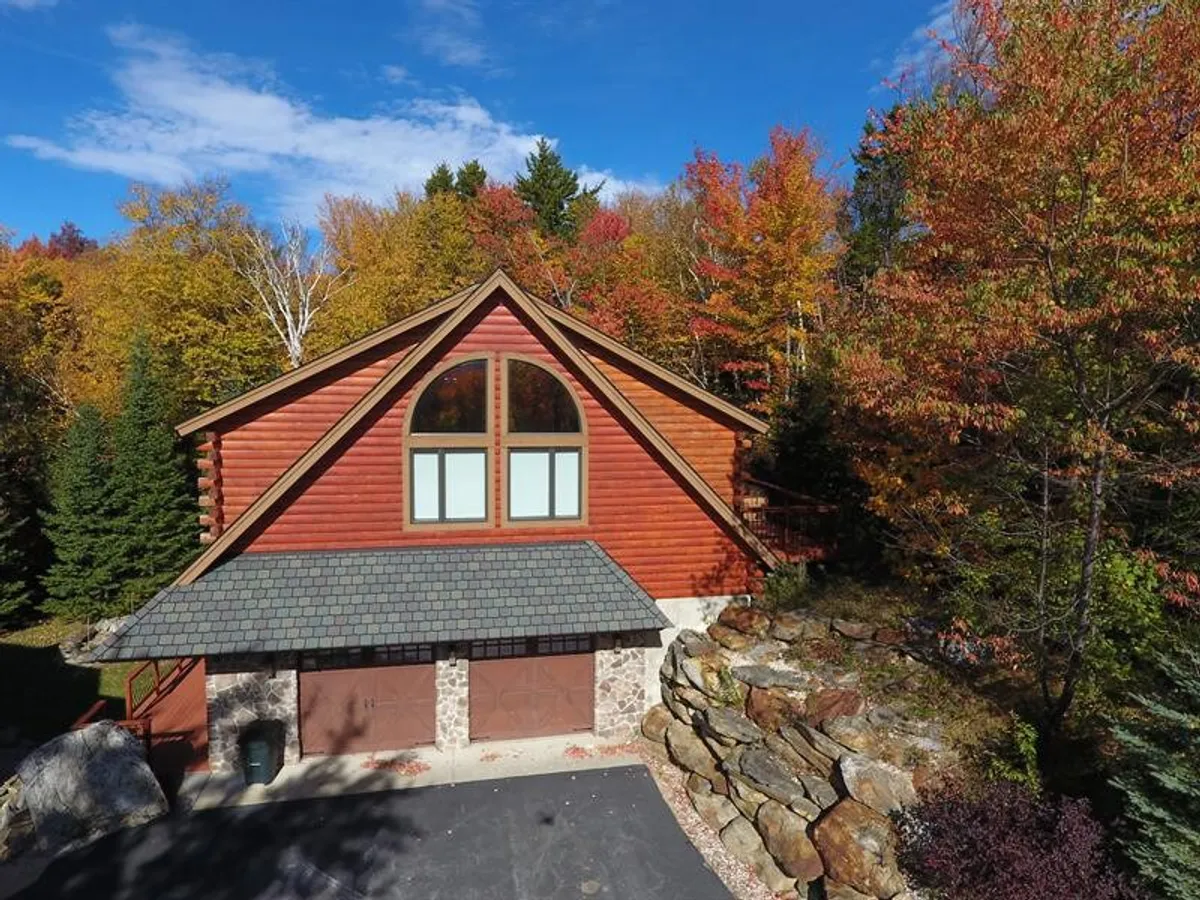 An exterior view of a log cabin structure with large arched windows and a deck surrounded by colorful autumn foliage. The scene suggests opportunities for outdoor barbecues, relaxing on the porch, and enjoying scenic mountain views from the deck area.