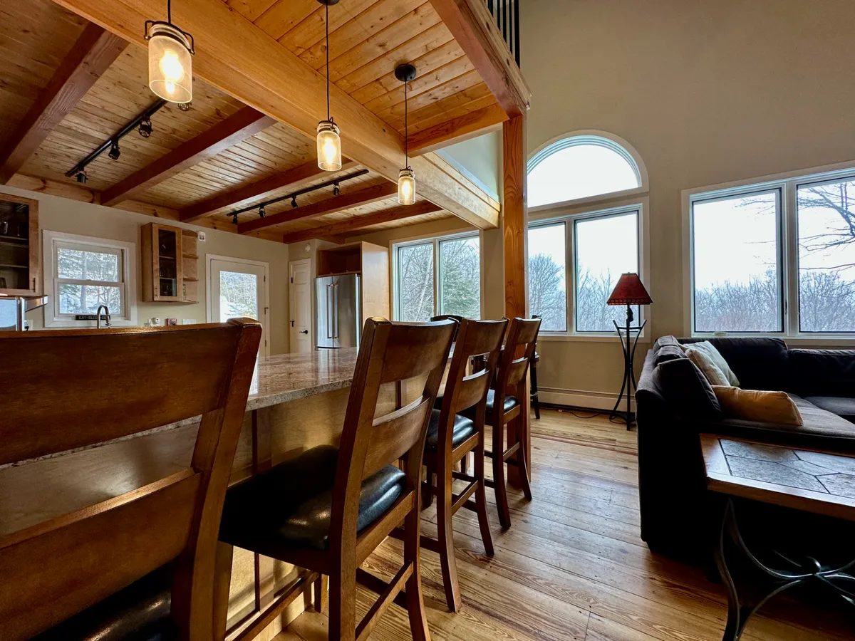 This image features a large wooden island with bar stools, a cozy sofa, and prominent large windows that invite natural light. The wooden beams across the ceiling add a warm, rustic charm, while the view through the windows hints at the outdoor scenery.