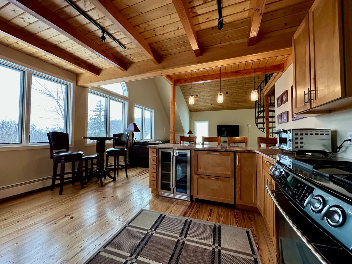 A spacious, modern kitchen featuring a prominent kitchen island surrounded by bar stools, large windows letting in natural light, and exposed wooden beams overhead. The space is arranged for both cooking and entertaining, with guests enjoying meals and socializing. Through the windows, sunset views provide a warm backdrop, enhancing the inviting mood.