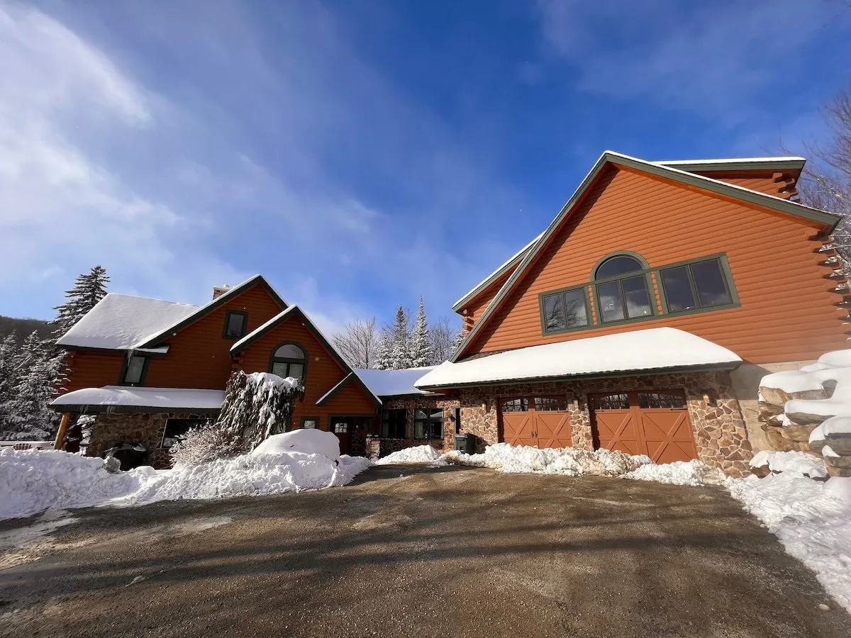 This exterior image captures the large stone entrance of the house with its distinct architectural style, set against a snow-covered driveway. The setting exudes warmth and a welcoming atmosphere, with hints of winter activities in the yard.