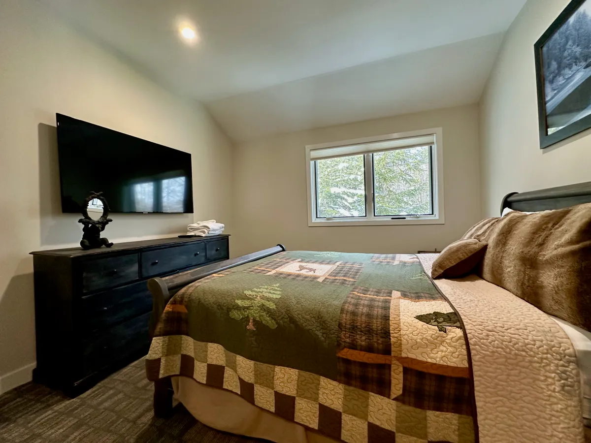 An inviting bedroom with a bed dressed in a patterned quilt, a window offering a view of trees, a dresser, and a television. The space feels warm and home-like, ideal for unwinding after a day of adventure.