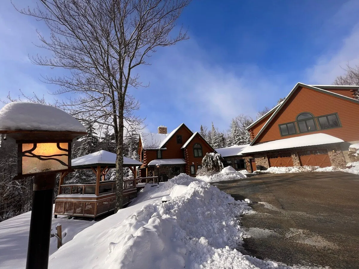 This exterior image features the main house nestled in a snowy landscape, with a cozy lamp post guiding the driveway. The scene evokes the atmosphere of winter sports and family gatherings, with visible opportunities for building snowmen or sipping hot cocoa on the porch. The snow-covered ground and inviting architecture emphasize the property's proximity to ski trails and its suitability for après-ski relaxation.