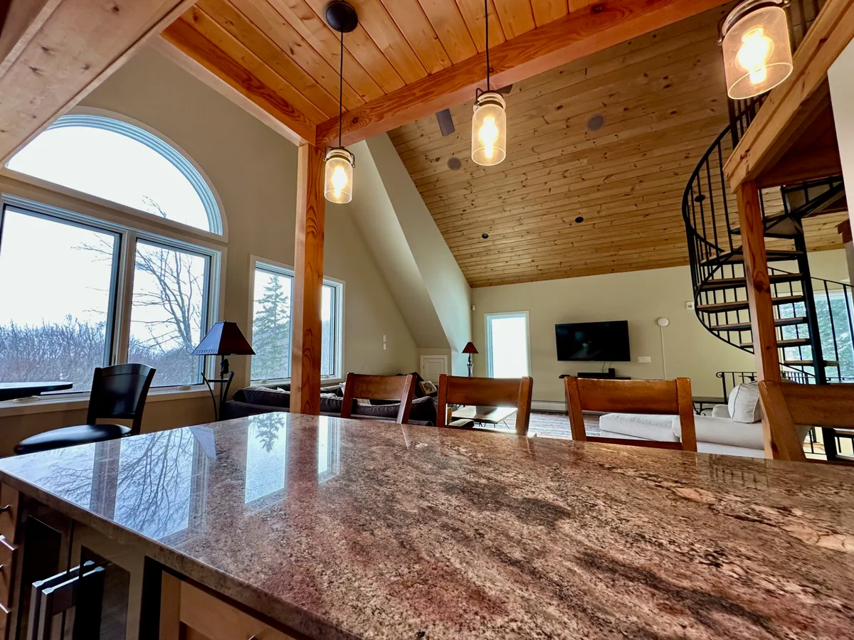 The image showcases a spacious open-plan living area with a granite countertop, spiral staircase, large windows offering scenic views, exposed wooden beams, and modern lighting fixtures. The living space looks inviting and is perfect for relaxing or socializing after a day spent outdoors.
