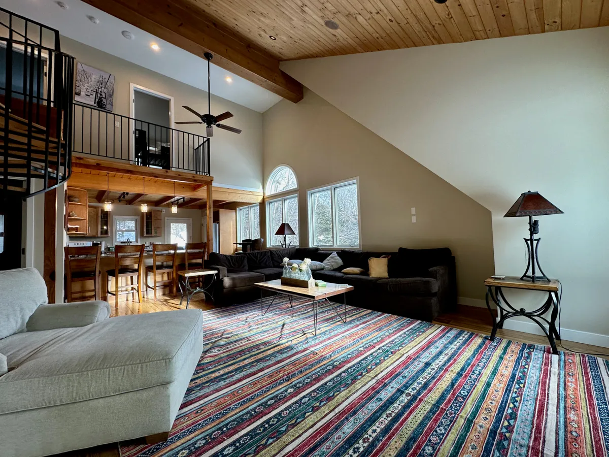 A comfortable, light-filled living space featuring a sofa, coffee table, spiral staircase, and expansive windows. The room is designed for relaxation, socializing, and enjoying both summer and winter days.