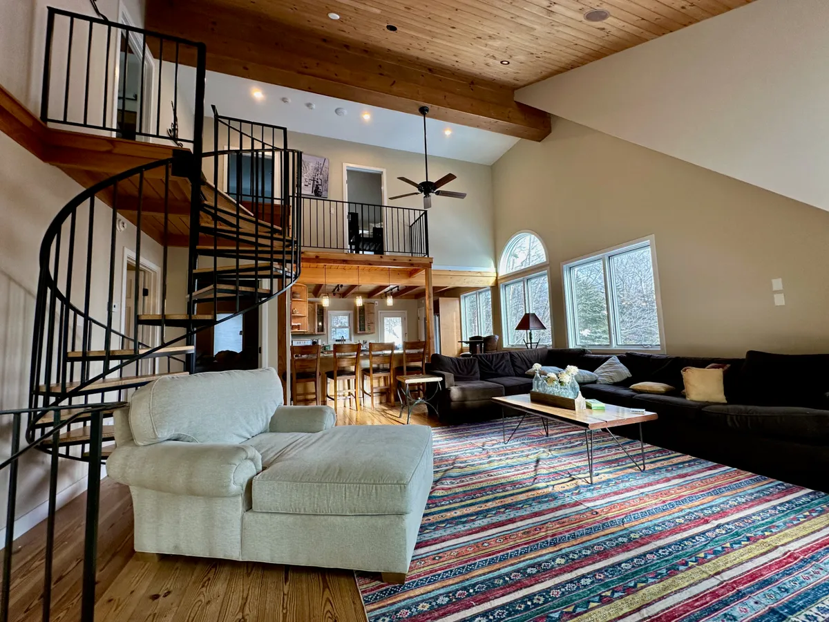 This interior image showcases a comfortable seating area beside a spiral staircase and large windows that frame the snowy outdoors. A colorful area rug and cozy seating create a welcoming environment for relaxing with family, entertaining guests, or enjoying mountain views after a day skiing. The windows visually connect the inside warmth to the adventure awaiting outside.