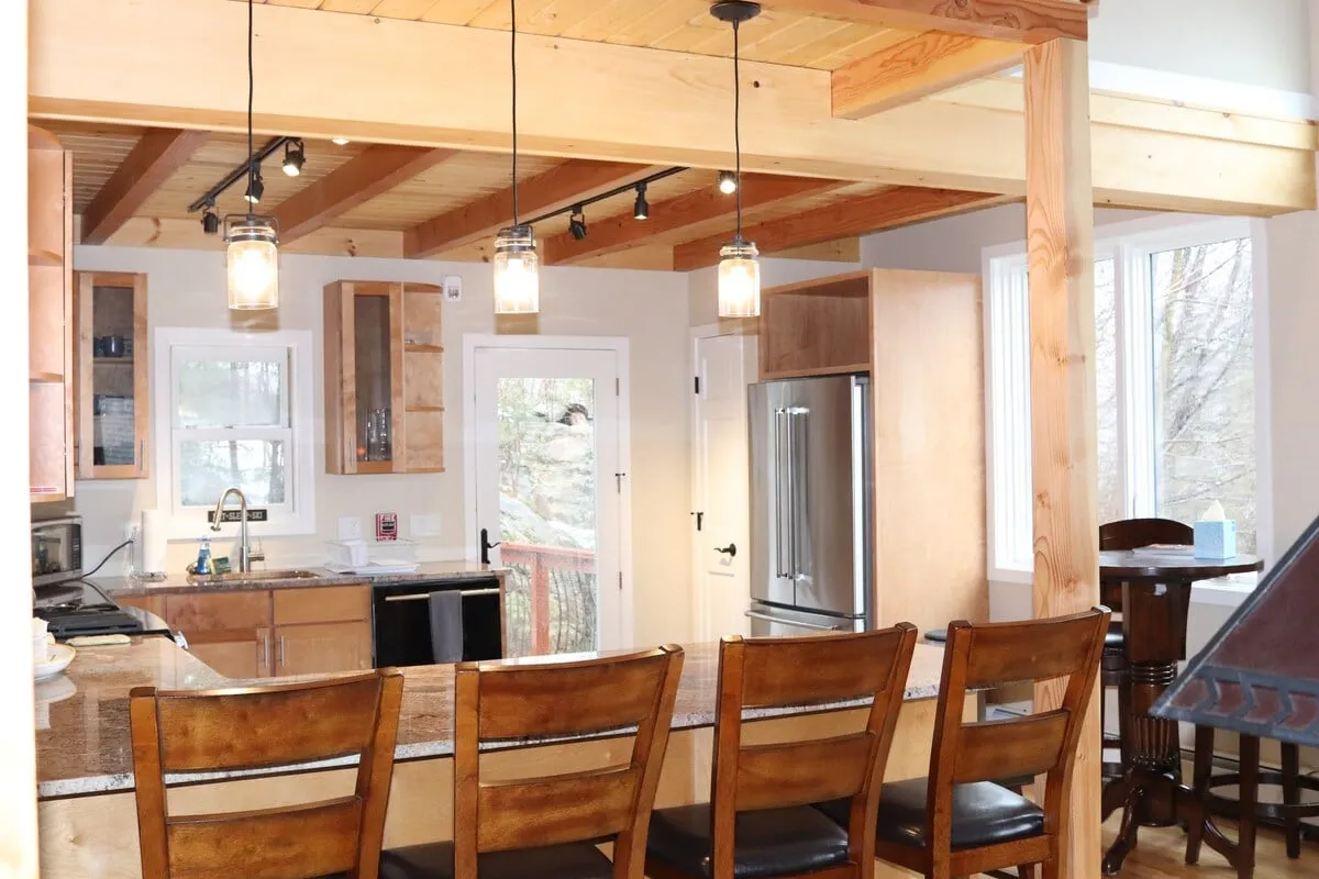 A bright, inviting kitchen featuring a kitchen island with bar stools, hanging light fixtures, and open windows that provide natural light. The scene easily accommodates several guests and highlights a setting perfect for cooking, brunches, or casual gatherings.